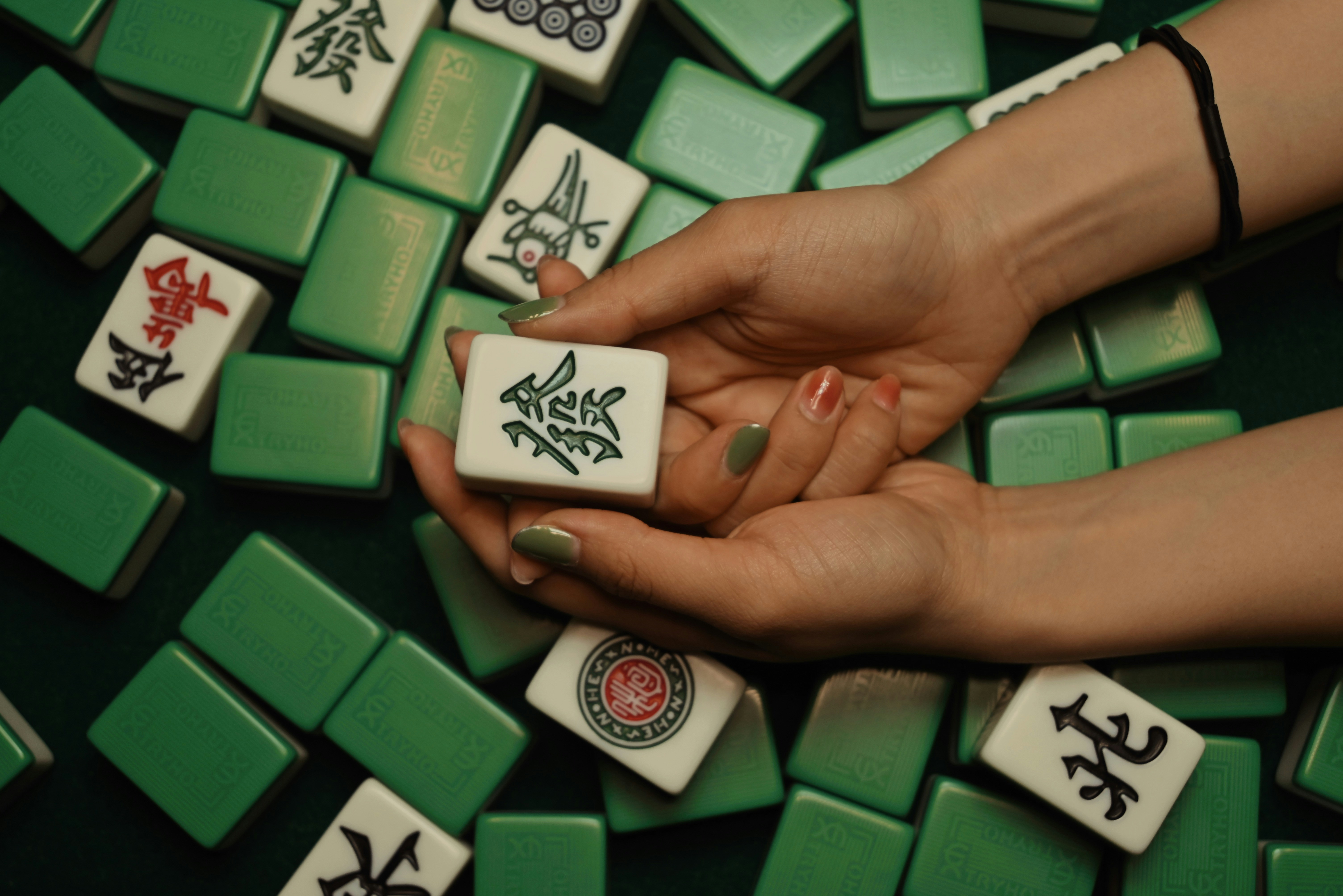 Hands holding green dragon tile above mahjong tiles. 
Photo by Ness fu on Unsplash