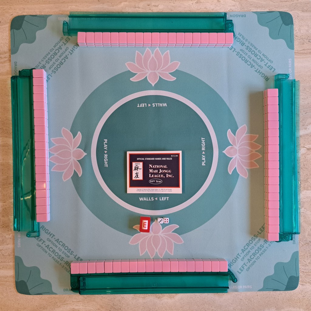 American Mahjong game setup with four green racks, pink Mahjong tiles arranged in 19-tile walls, National Mah Jongg League (NMJL) scorecard, wind indicator, and two dice on a game table.