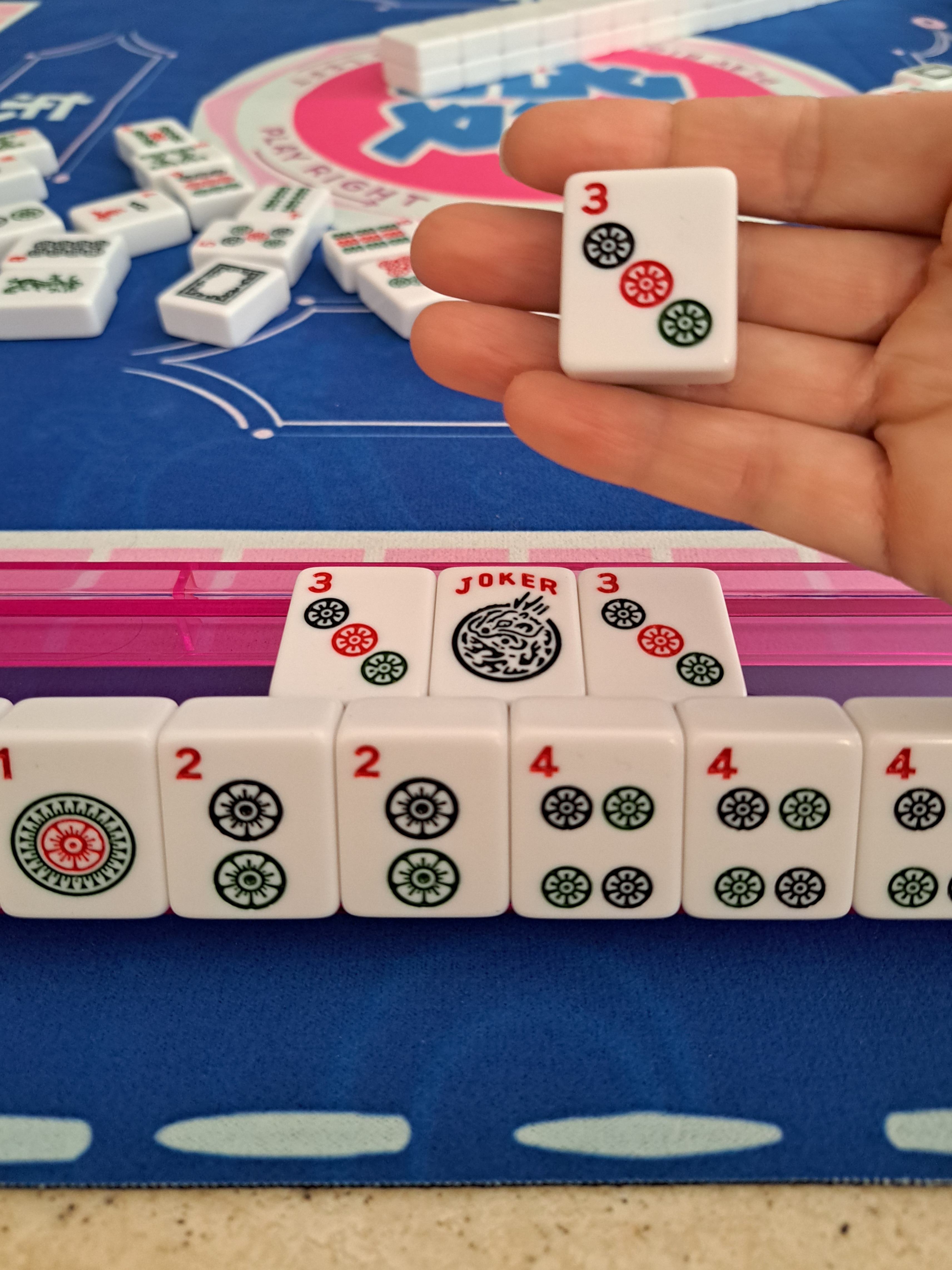 Mahjong player holding a newly drawn tile from the wall, preparing to exchange it for a joker in their own exposed Pung on the rack.