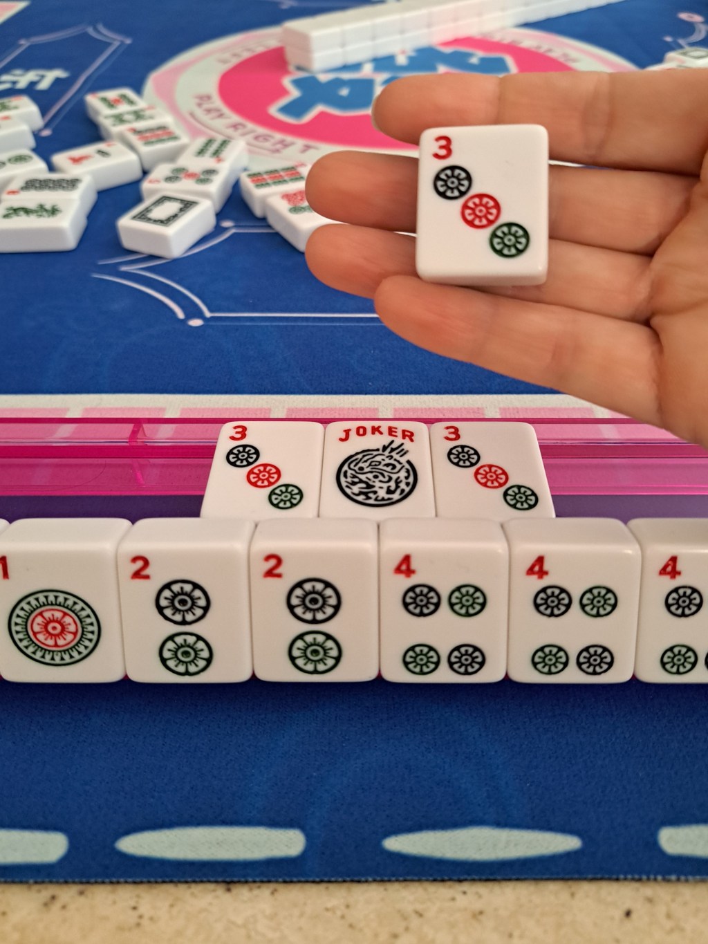 Mahjong player holding a newly drawn tile from the wall, preparing to exchange it for a joker in their own exposed Pung on the rack.