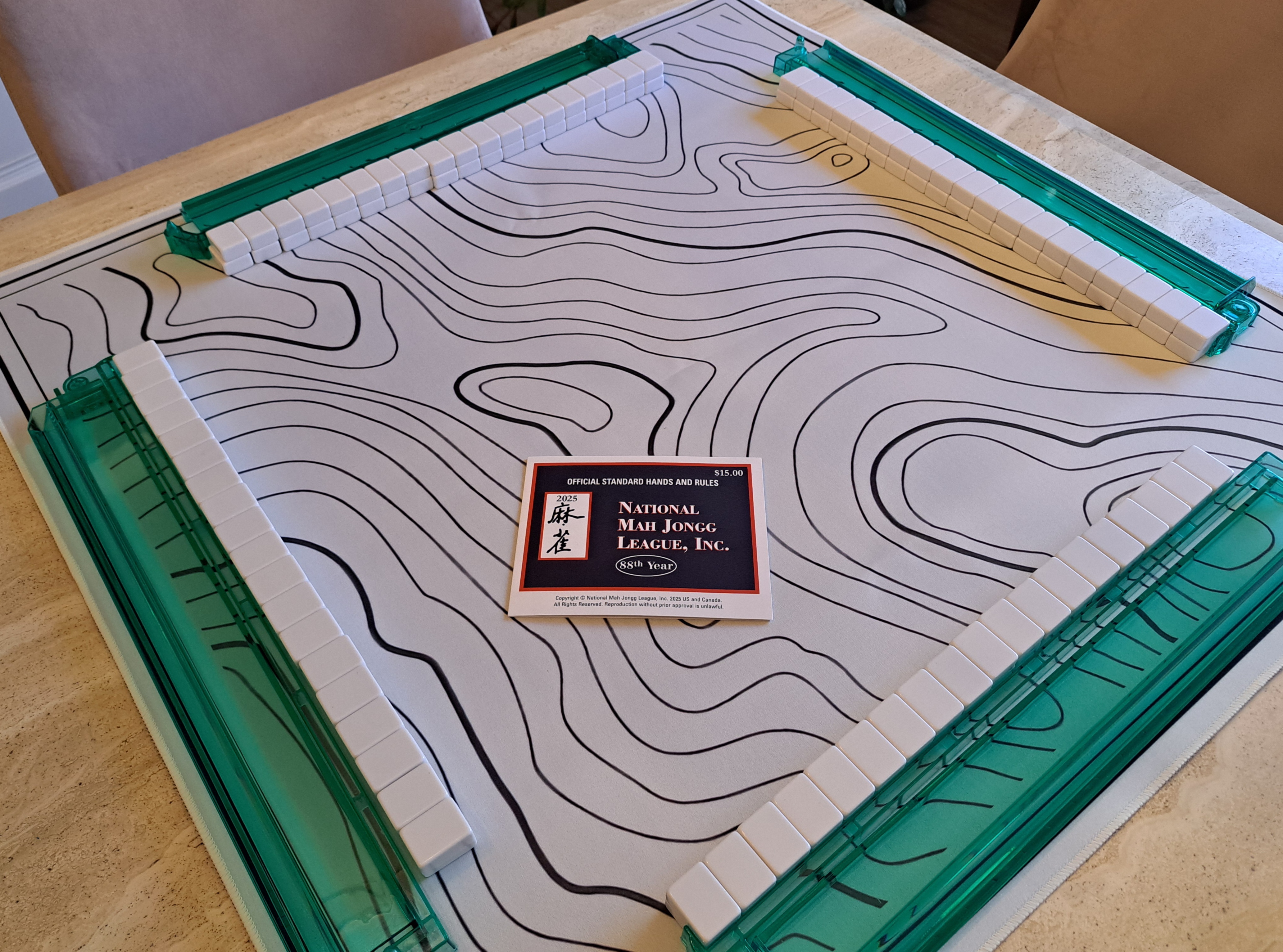 American Mahjong setup on a white mat with lined sections, four green racks holding walls of white tiles, and the 2025 National Mah Jongg League card displayed on the table.