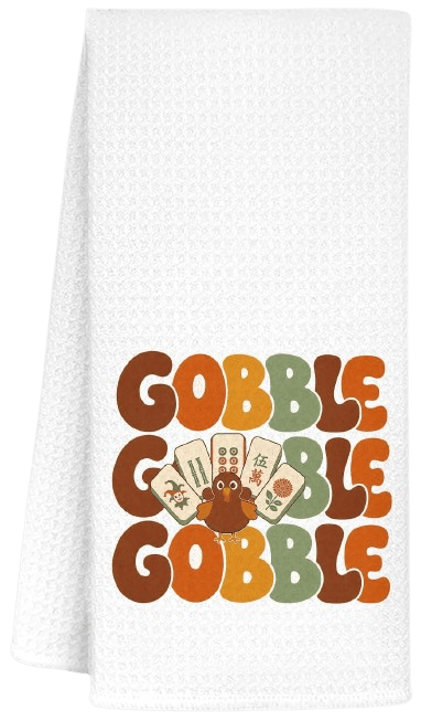 A festive kitchen towel with the words 'GOBBLE GOBBLE GOBBLE' in playful, colorful typography, featuring a cartoon turkey and various Thanksgiving-themed illustrations.
